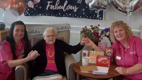 Betty Harrod with care workers Colette and Sharon at Aran Court Care Home. Ms Harrod is holding the other women's hands and is surrounded by birhtday cards and gifts as well as balloons and a sign saying "100 & fabulous".