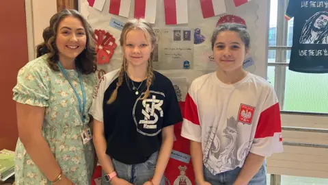 Three girls standing in front of a display for Poland. The girl on the left has light brown hair and wearing a green floral dress and lanyard. The girl in the middle has blonde hair in plaited pigtails and is wearing a black and white t-shirt, necklace and jeans. The girl on the right has brown hair tied back and is wearing a red and white graphic t shirt with Polish tag, and jeans.