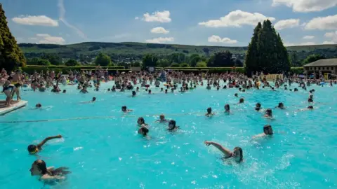 Children swim in an outdoor pool on a sunny day. There are moors visible in the background