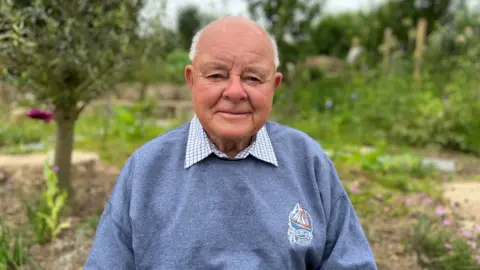 Viv Stratton is standing in the orchard as he smiles at the camera. He is wearing a checked shirt and a sweatshirt with an indistinct logo. Behind him are trees and flowers.