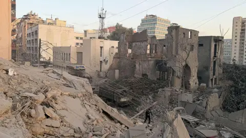 Ibrahim AMRO / AFP via Getty Images A pile of dusty rubble in central Beirut, surrounded by other buildings standing, as a man walks in the destruction.