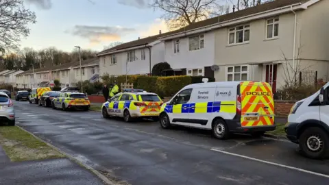 A row of cream and white semi-detached houses with two police vans and two police cars parked on the road outside, among other cars. Two police officers can be seen outside one of the houses 