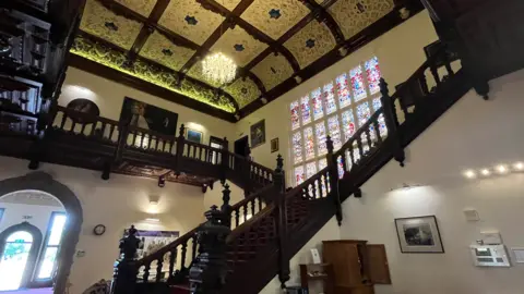 A large wooden stair case leads to a stained-glass window