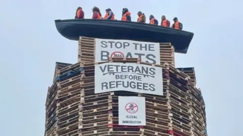 The picture shows a model of a small boat with figures inside on top of a tall bonfire constructed mainly from pallets. Signs saying "Stop The Boats" and "Veterans Before Refugees" are on the bonfire.