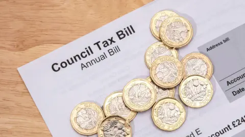 Getty Images A white piece of paper that reads "council tax bill, annual bill". There are twelve £1 coins sitting on top of it. The items are on a light wood table