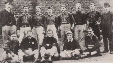 The black and white image shows two rows of footballers, one with them sitting and the other with the players standing. Some of the men have impressive mustaches. 