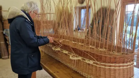 Supplied Felicity is wearing a blue coat and is standing at a table which has a half-built willow coffin on which she is weaving