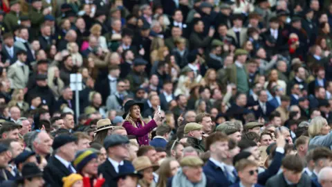 Image shows a huge crowd of people, all dressed formally, looking and pointing their phones in one direction. Image is focused on a woman wearing a purple jacket taking a picture on her phone. 