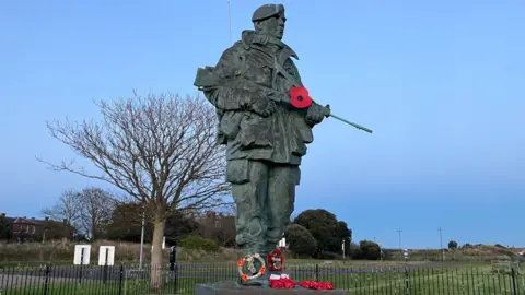 A Yomper statue outside a Royal Marines base in Southsea. It is a large blue statue of a man, carrying a gun and wearing full outdoor kit. There are poppy wreaths at his feet, and greenery in the background.