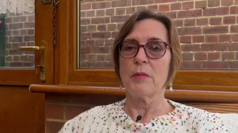 Chrissie Reidy/BBC A woman wearing glasses and a white woollen jumper with multi-coloured fleckles on it.