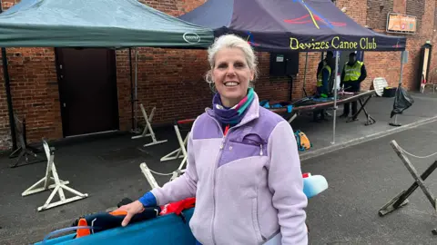 Emily King smiles at the camera - she is holding her paddleboard and wearing a purple fleece. Behind her, a red brick building and a gazebo in front, branded with the Devizes Canoe Club
