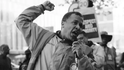 Supply/IWGB Union, a black and white picture, reflects a man, Helio Santos, which appears to be an opposition. He has a microphone in one hand and his other hand is raised in a fist. There are signs in the background, and one reads: "Honor and dignity"