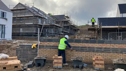 Housebuilders working on site in yellow high vis. There is a brick wall in the foreground and houses under construction in the background.