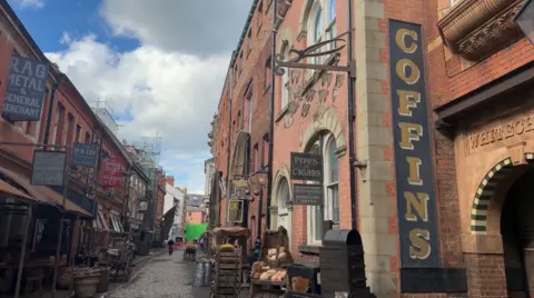 BBC News A cobbled street lined with Victorian warehouses and former shops and houses has been made up to look like Victorian London, with signs advertising "coffins", "pipes and cigars" and "rag metal", along with period street furniture.