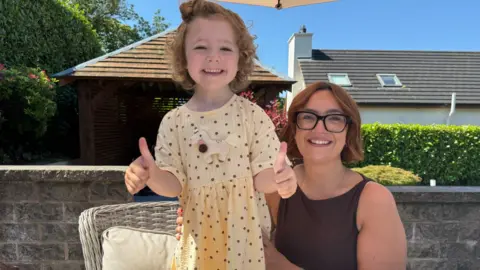 Hatty and her mother Victoria are outside under a bright blue sky. Hatty, who is wearing a yellow dress with purple polka dots, is standing up and smiling while holding her two thumbs up. Victoria is sitting down. She had short re/brown hair and is wearing a sleeveless dark purple top and black framed glasses. 