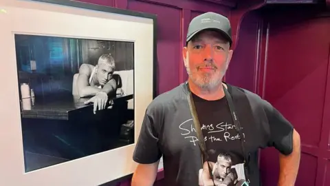Steve Read stands beside a framed black-and-white photograph. Read is wearing a dark baseball cap and a black T-shirt featuring artwork from the exhibition. The photograph beside him shows a blond-haired musician leaning on a counter, while burgundy-coloured wall panels form the background.
