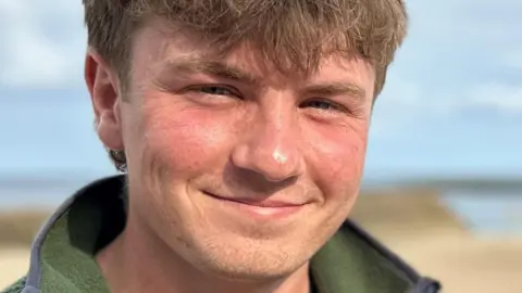 Euan and Andrew stand side by side smiling at the camera, with a yellow beach and blue sea behind them. Euan has brown hair and is wearing a green fleece. Andrew has black hair and is wearing a grey hoody beneath a navy body warmer.