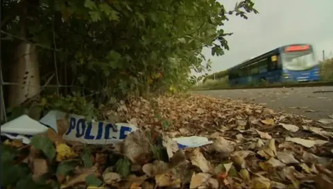 Image of police tape on the floor near to the busway, in and amongst leaves and bushes, with a bus going past in the background.