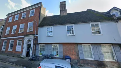 12 Mill Lane, Cambridge. it shows a two storey building with a front remodelled in the 18th Century to show four sash windows in the brick ground floor and three sash windows on the rendered first floor. To its right is a three storey Georgian brick building with rows of white sash windows. There is a pavement in front of both buildings. 