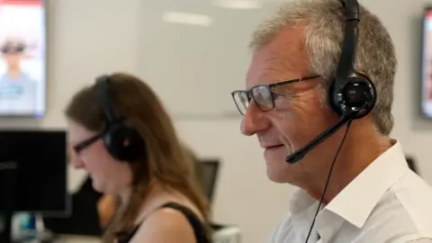 two call handlers can be seen, the man in the foreground is wearing a headset, a white shirt and has grey hair, a blurred woman in the background is wearing a headset and a black top