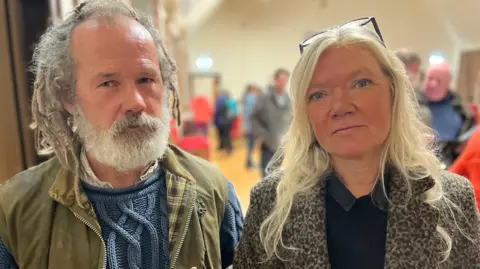 A bearded man with grey dreadlocks, wearing a green wax jacket a light shirt and a blue jumper and a blonde-hired woman wearing a leopard-print jacket and dark top with glasses on her head look at the camera. They are inside a hall with people in the background.