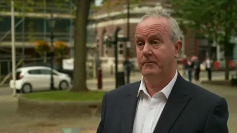 Martin Griffith in a suit jacket and white shirt. He is looking to the side of the camera. Behind him are trees, buildings and a white car.