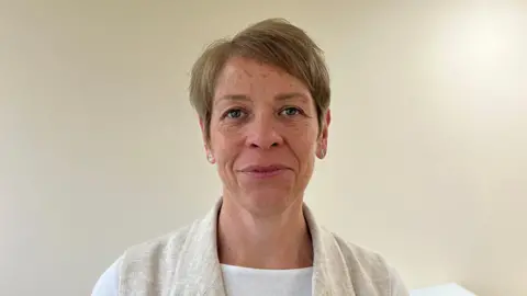 Short haired woman looks down the camera with a smirk. She is wearing a white T-shirt and a camel-coloured cardigan.