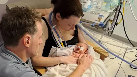 A mother and father are seen holding a baby in hospital. There are tubes around and going into the baby's nose and mouth.