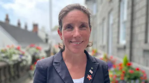 Deputy Lindsay de Sausmarez, who is wearing a blue jacket, with a white shirt. She has brown shoulder length hair. 