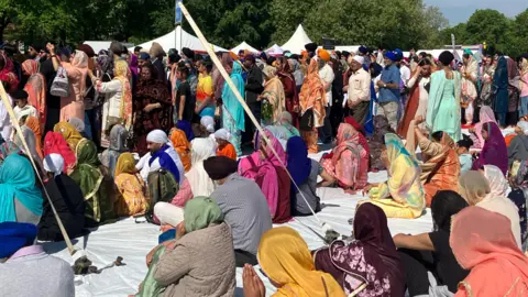 Sikh adults dressed in traditional clothing stand and sit on a white blanket in a park, facing a stage. Many have their hands together in prayer. White tent peaks can be seen behind, in front of trees.