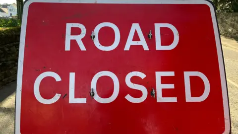 A Road Closed sign with white writing and a red background, a white border, zoomed in so you can only see parts of the road at the side of the sign.