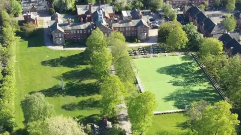 Cheshire Police An aerial view of an ornate building with a large field in front of it as well as a playing field and trees. There are rows of houses to the left hand side of the image.
