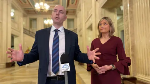 PA Media Jon Burrows is standing in front of a microphone to the left of the picture. To the left of the image is Diana Armstrong. They are standing in the Great Hall at Stormont addressing the press 