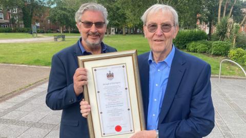 David Skentelbery and his son Gary, both of whom are grey-haired and wearing tinted glasses, blue blazers and blue shirts, hold a framed copy of the honorary freeman certificate