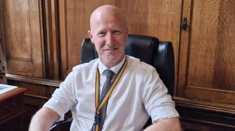 Lee Ratcliffe is wearing a white shirt with the sleeves rolled up and a blue and white patterned tie