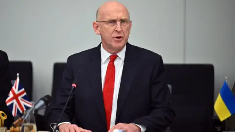 John Healey, who is bald and wears glasses, wears a black suit with a white shirt and red tie as he addresses a NATO meeting on Wednesday