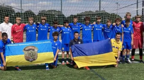 Teenage players from Ukraine stand and kneel in front of a goal with coaches either side of them. The four boys at the front are kneeling down and holding two flags with blue and yellow on them. The majority of the players are wearing a blue football kit and the two goalkeepers are wearing a red kit. The sun is shining and the sky is blue.