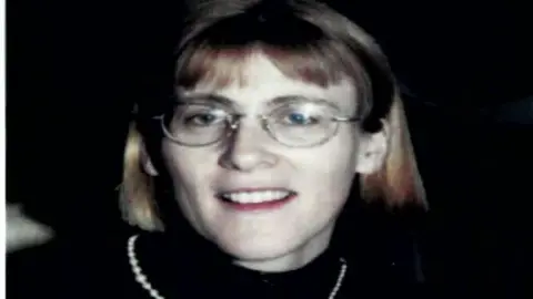 Family photo A family photo of the late Esther Gibson which was released to the Omagh Bombing Inquiry.  She has blonde hair and a fringe, and is wearing gold-rimmed glasses, a black top and a string of pearls.