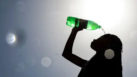 silhouette of a woman on the right side of the image pouring water over her head from a single use green plastic bottle. She is facing left and holding the bottle with her right hand. In the background is the glare of the sun