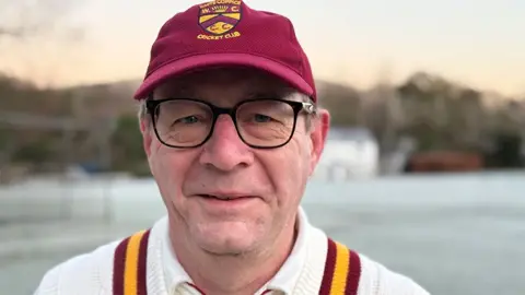 Paul O'Gorman/BBC Head and shoulders image of club chairman, Paul Dobson standing on a frost covered cricket pitch. He is wearing glasses, is smiling and is wearing cricket whites and a burgundy cap