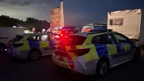 Two police cars with lights flashing parked behind a number of other vehicles with water and lights seen in the distance - it is dark.