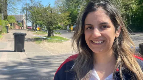 Toby Friedner/BBC A woman is smiling, behind her is police tape and a fire engine can be seen in the distance