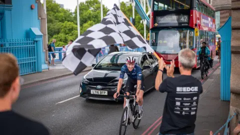 Rob Gardiner A woman rides on a bicycle towards us over a bridge, in front of a black car and red double decker bus. A man on the right is waving a black and white chequered flag.