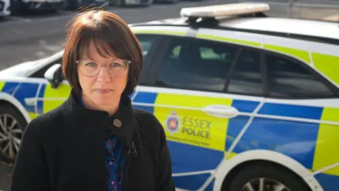 Jamie Niblock/BBC Date Ch Inspector Lewis Metcalfe with a dark bob haircut and glasses stands in front of a police car with the Essex Police logo and blue and yellow on it.