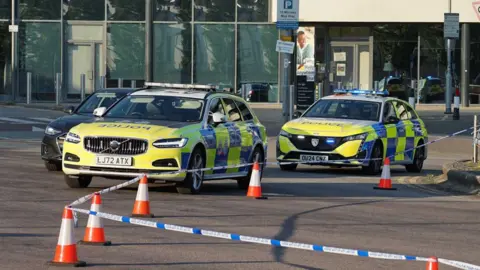 Police vehicles block off a section of road outside a modern glass-fronted building. Marked police cars are parked behind blue-and-white police tape and a line of orange traffic cones. Road signs, bollards and parked vehicles are visible around the cordon.