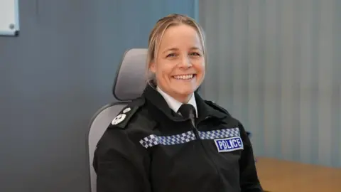 Gloucestershire Constabulary Katy Barrow-Grint, a young woman, sits in a chair at a desk in an office. She is wearing a black police jacket with a white shirt and black tie. She has blonde hair tied back.