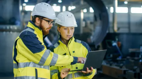 Getty Images Two engineers - one male, one female - wearing white hardhats and yellow hi-viz jackets standing in a factory and looking at a laptop