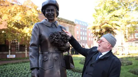 Reuters A man wearing a suit, glasses and a hat puts his right hand up to a bronze statue of Queen Elizabeth II.