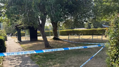 Police tape surrounding a number of trees, play equipment and basketball court