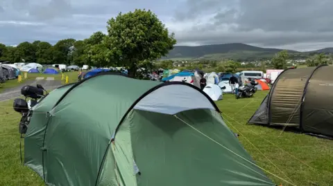 A field full of tents, two large green tents sit in foreground. You can also see some motorbikes parked up next to some tents further away.